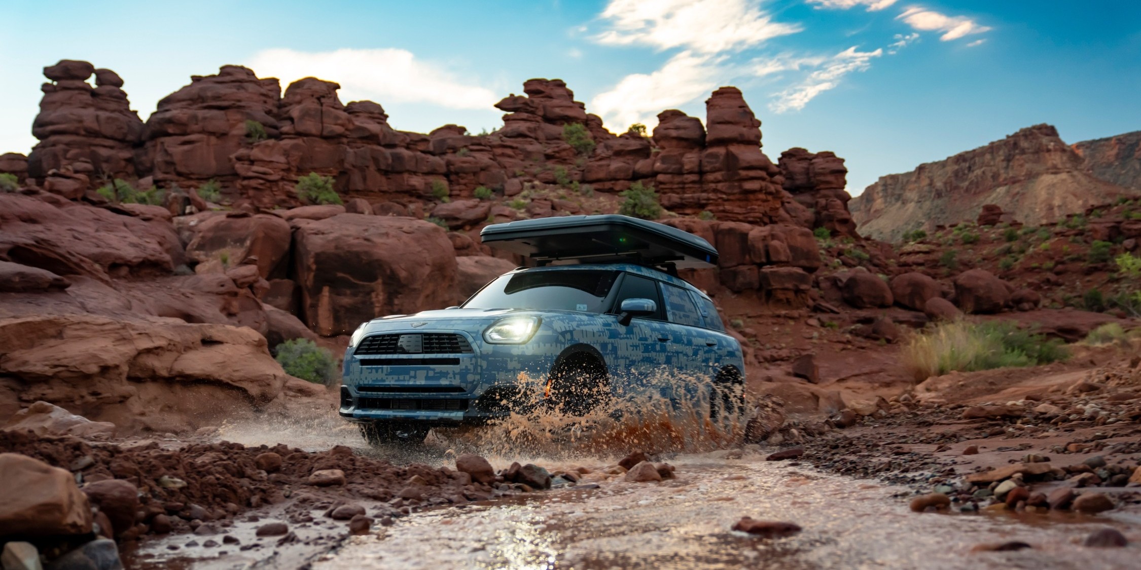 MINI Countryman a atravessar um riacho numa paisagem rochosa, com salpicos de  água e uma caixa de tejadilho 